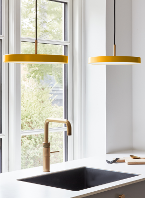 Asteria mini taklampe fra Umage i gul. Henger to stykker over en kjøkkenvask og foran et vindu. lys på