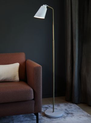 København gulvlampe i gullfarge fra Høvik lys med hvit skjerm stående ved en sofa, lys på