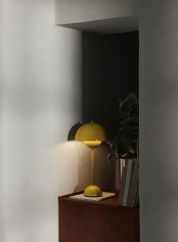 Flowerpot VP3 bordlampe H50 fra Tradition i gul. Plassert på en grå hylle ved siden av en seng. lys på