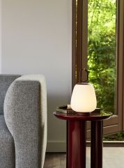 Lucca SC51 oppladbar bordlampe fra Tradition i rødbrun. Plassert på en overflate sammen med en plante og bøker stablet oppå hverandre. Lys på
