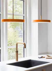 Asteria mini taklampe fra Umage i oransje. henger to stykker sammen over en kjøkkenvask og foran et vindu. lys på